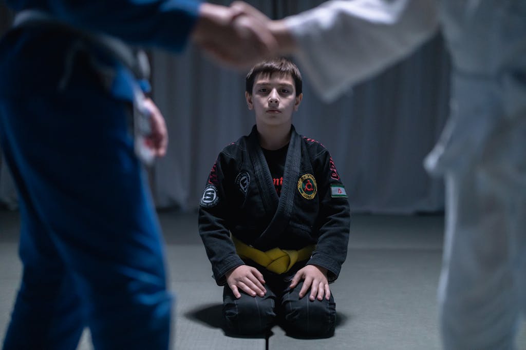 A young boy in a yellow belt gi kneels during a jiu jitsu training session indoors.